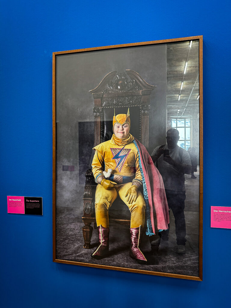 Ein Junge beziehungsweise junger Mann sitzt in einem gelben Superhelden-Anzug auf einem Stuhl, der einem Thron ähnelt. Er lächelt zufrieden.