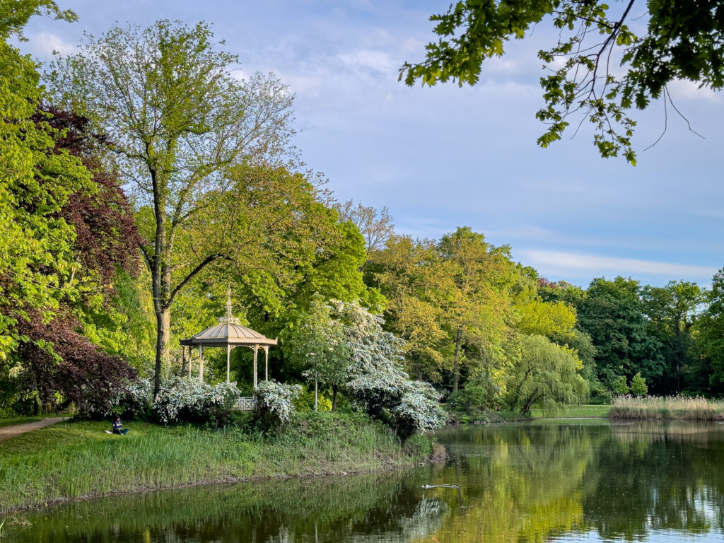 Ein Pavillon am grasbewachsenem Ufer ist umgeben von Büschen und Bäumen. Auf dem Rasen sitzt jemand. Rechts ist ein Teich zu sehen, dessen Ufer von Schilf und Bäumen gesäumt ist. Ins Bild ragen Äste mit grünen Blättern.