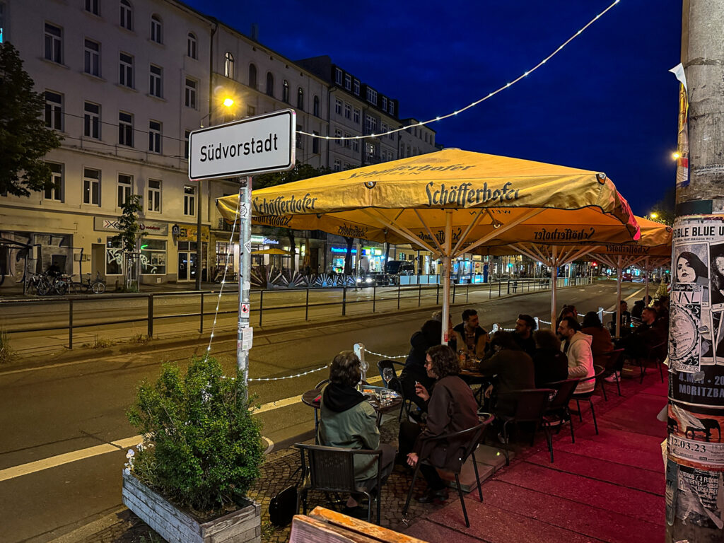 Vor der naTo – Café-Tische unter großen Sonnenschirmen laden zum Platz nehmen. Im Vordergrund ist ein Schild mit der Aufschrift Südvorstadt zu lesen, vor dem eine Pflanze steht. Dahinter liegt die Fahrbahn und die Gleise der Karl-Heine-Straße. Auf der gegenüberliegenden Straßenseite sind Wohnhäuser zu sehen.