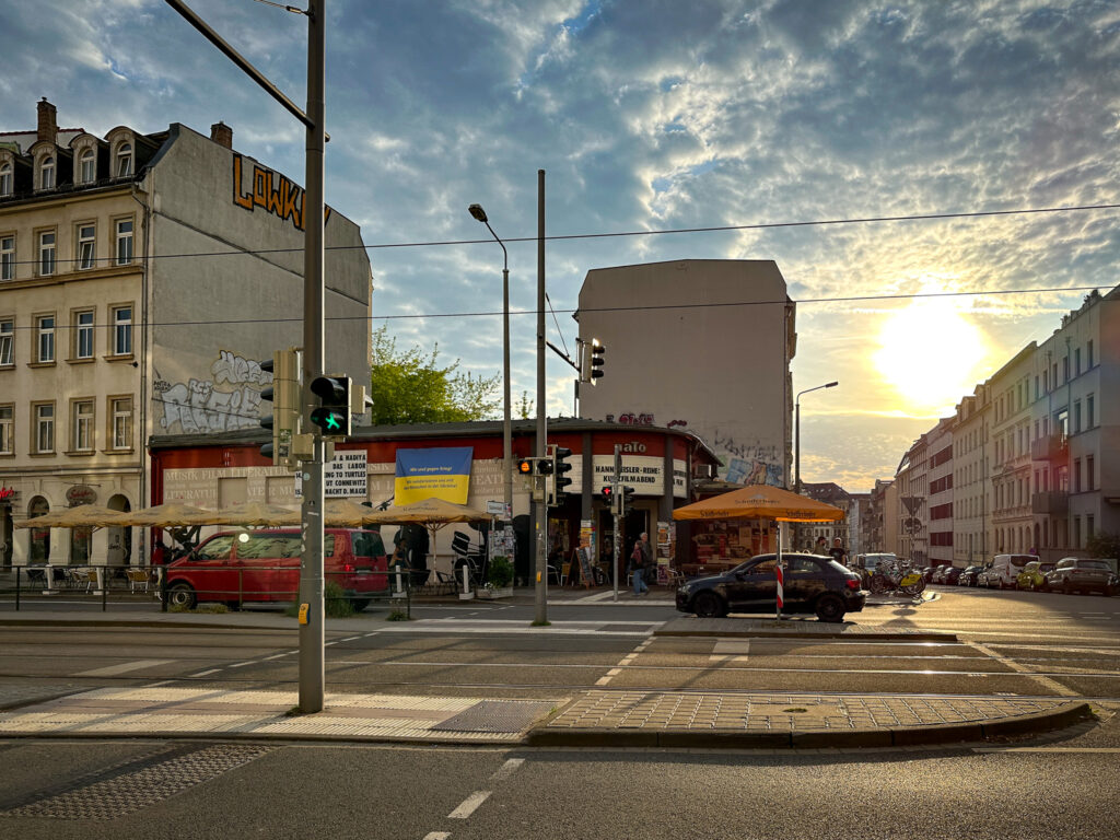Blick auf das Gebäude, das sich naTo und Cinémathèque teilen. Davor ist die Karl-Heine-Straße zu sehen. Zwei Autos fahren vorbei. Die im Vordergrund zu sehende Ampel des Fußgängerüberweges zeigt ein grünes Lichtzeichen. Der blaue Himmel ist voller Quellwolken, die von der gelben, untergehenden Sonne angestrahlt werden.